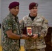 Two military personnel are jointly holding a commemorative plaque during what appears to be a formal ceremony. The soldier on the left wears a foreign military uniform with the name tag 'ASWAD' and an airborne patch, while the U.S. Army soldier on the right bears the name tag 'FAY' and wears the rank of Colonel (eagle insignia). A third soldier partially visible on the right has the name tag 'VARJIAN'.
