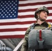A female military service member stands in front of a large American flag inside what appears to be a hangar or large military facility. She is wearing full combat/airborne gear including a tactical helmet, camouflage uniform (OCP pattern), and a parachute harness with reserve chute. The image is composed as a formal or promotional-style photograph with dramatic low-angle framing.