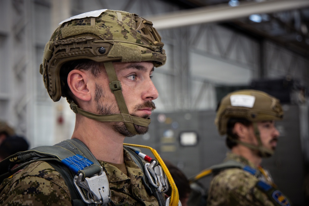 Two military personnel are shown in what appears to be a hangar or staging area, wearing tactical combat helmets and parachute harness equipment. The foreground subject is in sharp focus, displaying a digital camouflage uniform, a MICH/FAST-style helmet with chin strap, and visible parachute rigging including carabiners and colored deployment handles. The setting and equipment suggest pre-jump preparation for a parachute operation.