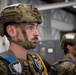 Two military personnel are shown in what appears to be a hangar or staging area, wearing tactical combat helmets and parachute harness equipment. The foreground subject is in sharp focus, displaying a digital camouflage uniform, a MICH/FAST-style helmet with chin strap, and visible parachute rigging including carabiners and colored deployment handles. The setting and equipment suggest pre-jump preparation for a parachute operation.