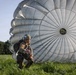 A military paratrooper has just landed in a grassy field with a large gray round parachute still inflated behind them. The soldier is in a crouched position, wearing full combat gear including a helmet with goggles and a backpack, and appears to be managing the parachute lines after touchdown. The setting appears to be a training exercise or operational jump at a drop zone surrounded by trees.