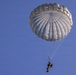 A military paratrooper is descending under a fully deployed round parachute against a clear blue sky. The parachute appears to be a standard military-issue T-10 or similar round canopy in off-white/gray color. The jumper is wearing full combat gear and appears to be in a controlled descent position.