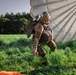 A military paratrooper in full combat gear has just landed in a grassy field, with their parachute still deployed and billowing behind them. The soldier is wearing multicam camouflage and a helmet bearing both a US flag patch and a Brazilian flag patch, suggesting a joint or multinational military exercise. An orange marker panel is visible on the ground in the foreground.
