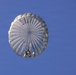 A parachutist is descending under a large, round, light gray/white military-style parachute against a clear blue sky. The image is taken from a low angle looking upward, providing a view of the parachute canopy from below. The parachutist appears to be wearing military or tactical gear.
