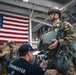A military parachute preparation scene inside a hangar, showing personnel being fitted and inspected with parachute equipment. A person wearing a 'JUMPMASTER' shirt is checking the gear of a soldier wearing Canadian flag patches on their uniform. A large American flag is prominently displayed in the background.