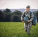 A smiling female soldier in full camouflage uniform walks across an open green field carrying military gear. She is wearing a combat helmet and carrying what appears to be a packed parachute or sleeping bag along with a large duffel bag. The image is taken during golden hour with a backdrop of trees and rolling hills.