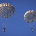 Two military paratroopers are descending under fully deployed round parachutes against a clear blue sky. Both parachutes are silver/gray in color and appear to be standard military static-line style canopies. The jumpers are wearing full military gear and appear to be in controlled descent.