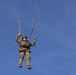 A military paratrooper is descending under a parachute canopy against a clear blue sky. The individual is wearing full military camouflage uniform and equipment, actively gripping the parachute risers/toggles to control descent. The parachute canopy itself is out of frame above, with multiple suspension lines visible converging upward.