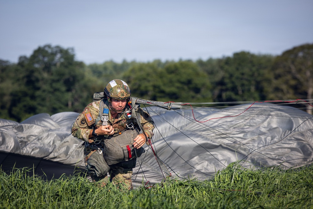 A U.S. Army soldier has just landed after a parachute jump and is crouching in a grassy field managing their parachute lines. The soldier is wearing full combat gear including a camouflage helmet, tactical vest, and harness, with a U.S. flag patch visible on the left shoulder. The deployed parachute canopy is spread out on the ground behind them.