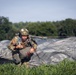 A U.S. Army soldier has just landed after a parachute jump and is crouching in a grassy field managing their parachute lines. The soldier is wearing full combat gear including a camouflage helmet, tactical vest, and harness, with a U.S. flag patch visible on the left shoulder. The deployed parachute canopy is spread out on the ground behind them.