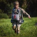 A soldier in multicam camouflage uniform and helmet is running across a grassy field while carrying parachute equipment, appearing to smile. A small flag patch (red, white, and blue — consistent with Netherlands/Luxembourg colors) is visible on the uniform. A second figure in camouflage is visible in the background, out of focus.