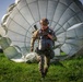 A military paratrooper has just landed in a green grassy field, with a large olive/grey round parachute fully deployed and spread out on the ground behind them. The soldier is wearing full combat gear including a helmet, camouflage uniform, and harness, and appears to be smiling or grimacing as they walk forward. The image is taken at low angle during what appears to be golden hour lighting conditions.