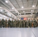 A large group of military personnel in camouflage uniforms are assembled inside what appears to be a military hangar or large indoor facility. An American flag is prominently displayed in the background, suggesting this may be a formal gathering, briefing, or deployment ceremony. The personnel are standing in a loose formation facing toward the right side of the frame.