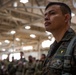 A military service member in digital camouflage uniform stands at attention inside a large hangar or military facility. The soldier wears visible patches including what appears to be a parachutist badge and unit insignia. A large formation of other uniformed personnel is visible in the blurred background.