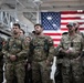 A group of military personnel in camouflage uniforms are assembled inside what appears to be a military hangar or large indoor facility. Several individuals are wearing Canadian Armed Forces uniforms with visible Canadian flag patches, while at least one person appears to be in a different camouflage pattern. A large American flag is prominently displayed on the wall in the background.
