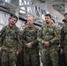 A group of military personnel in camouflage uniforms are standing at attention or in a relaxed formation inside what appears to be a military hangar or large indoor facility. Several individuals are visibly wearing Canadian Armed Forces patches and parachute harnesses, suggesting a joint military exercise or briefing. A large American flag is visible in the background, indicating a possible joint US-Canada military operation or event.
