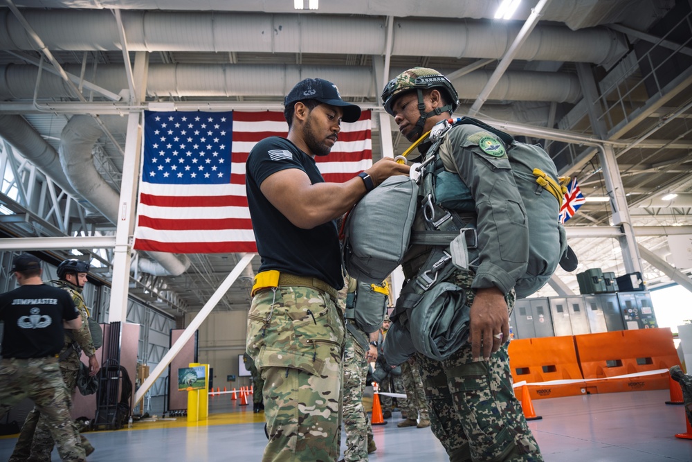 A military jumpmaster in a black t-shirt and camouflage pants is conducting a parachute equipment inspection on a soldier wearing full jump gear including a helmet and parachute pack. The setting appears to be a military hangar with a large American flag and a partially visible British flag in the background, suggesting a multinational military exercise or training event. Additional personnel in military uniforms are visible in the background.