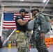 A military jumpmaster in a black t-shirt and camouflage pants is conducting a parachute equipment inspection on a soldier wearing full jump gear including a helmet and parachute pack. The setting appears to be a military hangar with a large American flag and a partially visible British flag in the background, suggesting a multinational military exercise or training event. Additional personnel in military uniforms are visible in the background.