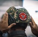 A close-up rear view of a person wearing a military tactical helmet covered in digital camouflage fabric. The helmet bears a red circular patch reading '10 BRIGED PARA' and 'TINDAK PANTAS', indicating affiliation with the Malaysian Army's 10th Parachute Brigade. The person's hands are adjusting the helmet, and a tactical headset is mounted on the side.