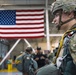 A U.S. Army Airborne soldier in full parachute jump gear stands in the foreground of what appears to be a military hangar. The soldier is wearing MultiCam (OCP) camouflage, a combat helmet, and is rigged with a parachute harness and static line. A large American flag is prominently displayed in the background, with other personnel visible in the hangar.