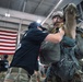 A military jumpmaster is conducting a pre-jump equipment inspection on a fully geared paratrooper inside what appears to be a military hangar. The jumpmaster, wearing a black 'JUMPMASTER' shirt and cap, is checking the parachute harness and rigging of the soldier, who is wearing full combat gear including a helmet and loaded parachute pack. An American flag is prominently displayed in the background, and additional personnel in military uniforms are visible throughout the hangar.