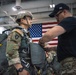 A U.S. Army Airborne soldier in full combat uniform and parachute harness is being inspected or fitted by an instructor in a military hangar. The soldier wears a helmet numbered '51' and displays Airborne and unit patches on his uniform. An American flag is prominently displayed on the hangar wall in the background.