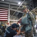 A military parachute equipment check is taking place inside a large hangar facility. A person wearing a black 'Jumpmaster' t-shirt is inspecting the parachute rig of a soldier in full combat gear and tactical helmet. Multiple other military personnel in camouflage uniforms are visible in the background, with a large American flag prominently displayed on the hangar wall.