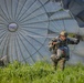 Two military paratroopers are shown immediately after landing in a grassy field, managing a large deployed parachute. The foreground soldier is actively pulling on the parachute risers/lines to collapse the canopy, while a second soldier is crouched low behind him. The large gray round parachute canopy remains partially inflated and dominates the background of the image.