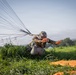 A military paratrooper has just landed in a grassy field and is crouching low while managing their parachute canopy and suspension lines. The individual is wearing full military gear including a tactical helmet, goggles, camouflage uniform, and a harness with a reserve parachute container. An orange marker panel is visible on the ground nearby, likely used as a landing zone indicator.
