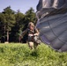 A military paratrooper has just landed in a green grassy field and is moving away from a large gray parachute that is still partially inflated behind them. The soldier is wearing full combat gear including a helmet, camouflage uniform, and harness with equipment attached. Additional personnel and equipment are visible in the background near a tree line.