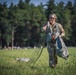 A smiling military paratrooper in camouflage uniform and tactical helmet is gathering and carrying a deployed parachute across a grassy field after a successful landing. The soldier appears in good spirits, holding the parachute lines and canopy bundle in both hands. The setting is an open drop zone surrounded by dense green trees under a clear sky.