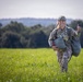 A female soldier in full camouflage uniform walks across a green field while giving a thumbs-up gesture and smiling. She is carrying what appears to be a packed parachute or large equipment bag, along with a backpack, and is wearing a military helmet. The setting appears to be a rural drop zone or training area with trees and hills visible in the background.