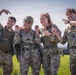 Four individuals wearing British Army MultiTerrain Pattern (MTP) camouflage uniforms are posing cheerfully in an open green field. They appear to be in a celebratory mood, smiling and gesturing toward one another. The setting and equipment suggest a military training or parachuting exercise has just been completed.