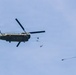 A military CH-47 Chinook tandem-rotor helicopter is airborne against a pale blue sky, with multiple parachute bundles or cargo drops visible trailing behind and below it. At least two separate parachute deployments are visible — one near the helicopter and one further to the right at a lower altitude. This appears to be a military airdrop or resupply exercise.