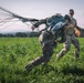 Two military personnel are visible in an open grassy field, with one individual managing a deployed parachute and its suspension lines after an apparent landing. The parachutist is bent forward, struggling to control the parachute canopy being pulled by wind, while a second person in camouflage uniform and baseball cap holds a clipboard nearby. The scene appears to be a military airborne training or operational jump exercise.