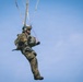 A military paratrooper is descending under a parachute against a clear blue sky. The soldier is wearing full combat gear including camouflage uniform, helmet, and a large tactical backpack. The image captures the soldier mid-descent, holding parachute control lines.