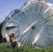 A military paratrooper has just landed on a grassy field with a large round parachute still partially inflated behind them. The soldier is wearing full combat gear including a helmet, sunglasses, camouflage uniform, and a large rucksack. The image captures the immediate post-landing moment with the canopy billowing in the wind.