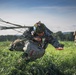 A military paratrooper is captured at the moment of landing in a green field, being dragged forward by parachute lines still attached and billowing behind them. The soldier is in full combat gear including helmet, tactical vest, and equipment pack, struggling to control the parachute upon touchdown. Additional personnel in military uniforms are visible in the background of what appears to be a training or operational jump exercise.