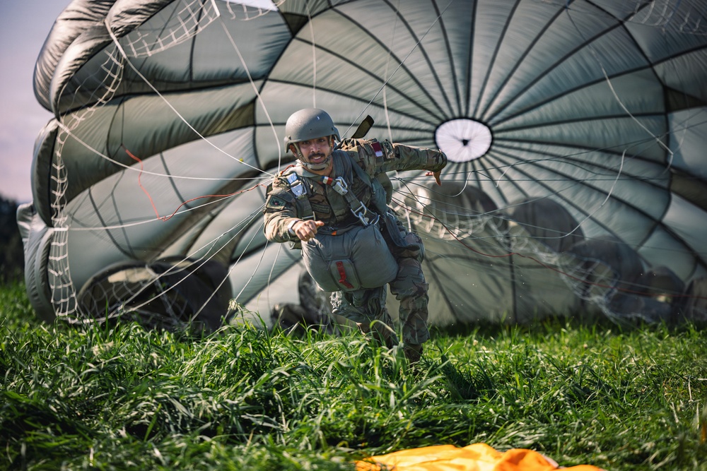 A military paratrooper has just landed on a grassy field and is actively managing a large olive-drab round parachute that is still partially inflated behind him. The soldier is wearing full combat gear including a helmet, tactical vest, and harness, and appears to be pulling on the parachute lines to collapse the canopy. An orange object, likely a deployment bag or marker, is visible on the ground in the foreground.