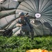 A military paratrooper has just landed on a grassy field and is actively managing a large olive-drab round parachute that is still partially inflated behind him. The soldier is wearing full combat gear including a helmet, tactical vest, and harness, and appears to be pulling on the parachute lines to collapse the canopy. An orange object, likely a deployment bag or marker, is visible on the ground in the foreground.