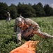 A soldier in full camouflage uniform and tactical helmet is on the ground in a grassy field, gathering and securing parachute equipment after a landing. An orange parachute canopy is visible on the ground in front of the soldier, along with suspension lines. Additional personnel and parachute equipment are visible in the background.
