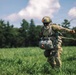 A military paratrooper has just landed in a green grassy field and is managing their parachute lines after touchdown. The individual is wearing full tactical gear including a camouflage uniform, helmet with visor, sunglasses, harness, and a pack. The parachute canopy lines are still visible trailing behind them as they move across the landing zone.