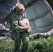 A military paratrooper in camouflage uniform is standing in a grassy field after a parachute landing, gathering and bundling his parachute canopy. He is wearing full jump gear including a tactical helmet, harness, and carrying equipment. A large deployed parachute canopy is visible in the background, partially collapsed on the ground.