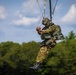 A military paratrooper is suspended in mid-air, appearing to be during a parachute descent or training exercise. The soldier is wearing full camouflage gear, a helmet, and is connected to suspension lines visible above. Green trees and a partly cloudy sky form the background.