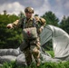 A U.S. Army soldier in full combat gear and parachute harness has just landed in a grassy field, with a deployed parachute canopy visible on the ground behind them. The soldier appears to be recovering from landing, moving forward with arms outstretched for balance. The setting appears to be a designated drop zone surrounded by trees under a partly cloudy sky.