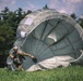 A military paratrooper has just landed on a grassy field and is managing a large silver-gray round parachute that is still partially inflated and billowing on the ground. The soldier is wearing full combat gear including a helmet and camouflage uniform, and appears to be pulling on the parachute lines to collapse the canopy. The scene is set against a backdrop of dense evergreen trees under a partly cloudy sky.