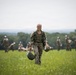 A smiling female soldier in camouflage uniform walks across a green field carrying a helmet and what appears to be a packed parachute or kit bag. She is in sharp focus in the foreground while multiple other military personnel in similar uniforms are visible in the blurred background. The setting appears to be a military training exercise or parachute landing field during summer conditions.