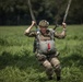 A military paratrooper in full combat gear is captured at the moment of landing in a grassy field. The individual is wearing multicam camouflage uniform and helmet, gripping parachute risers with both hands in a landing posture with knees bent. The background shows a green meadow with dense trees, suggesting a military training or operational jump exercise.