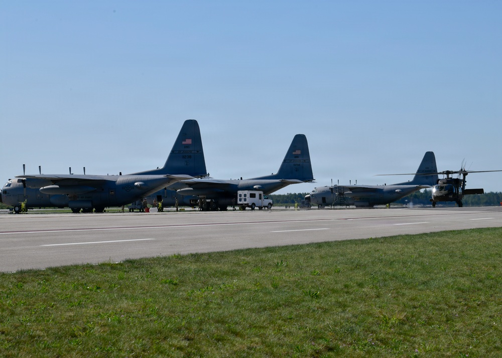 The image shows a military flight line with multiple large transport aircraft and a helicopter parked on a tarmac. At least two C-130 Hercules turboprop aircraft are prominently visible alongside what appears to be a UH-60 Black Hawk helicopter on the right side. Ground crew personnel and support vehicles are visible between the aircraft, suggesting active maintenance or pre-flight operations.
