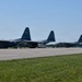 The image shows a military flight line with multiple large transport aircraft and a helicopter parked on a tarmac. At least two C-130 Hercules turboprop aircraft are prominently visible alongside what appears to be a UH-60 Black Hawk helicopter on the right side. Ground crew personnel and support vehicles are visible between the aircraft, suggesting active maintenance or pre-flight operations.